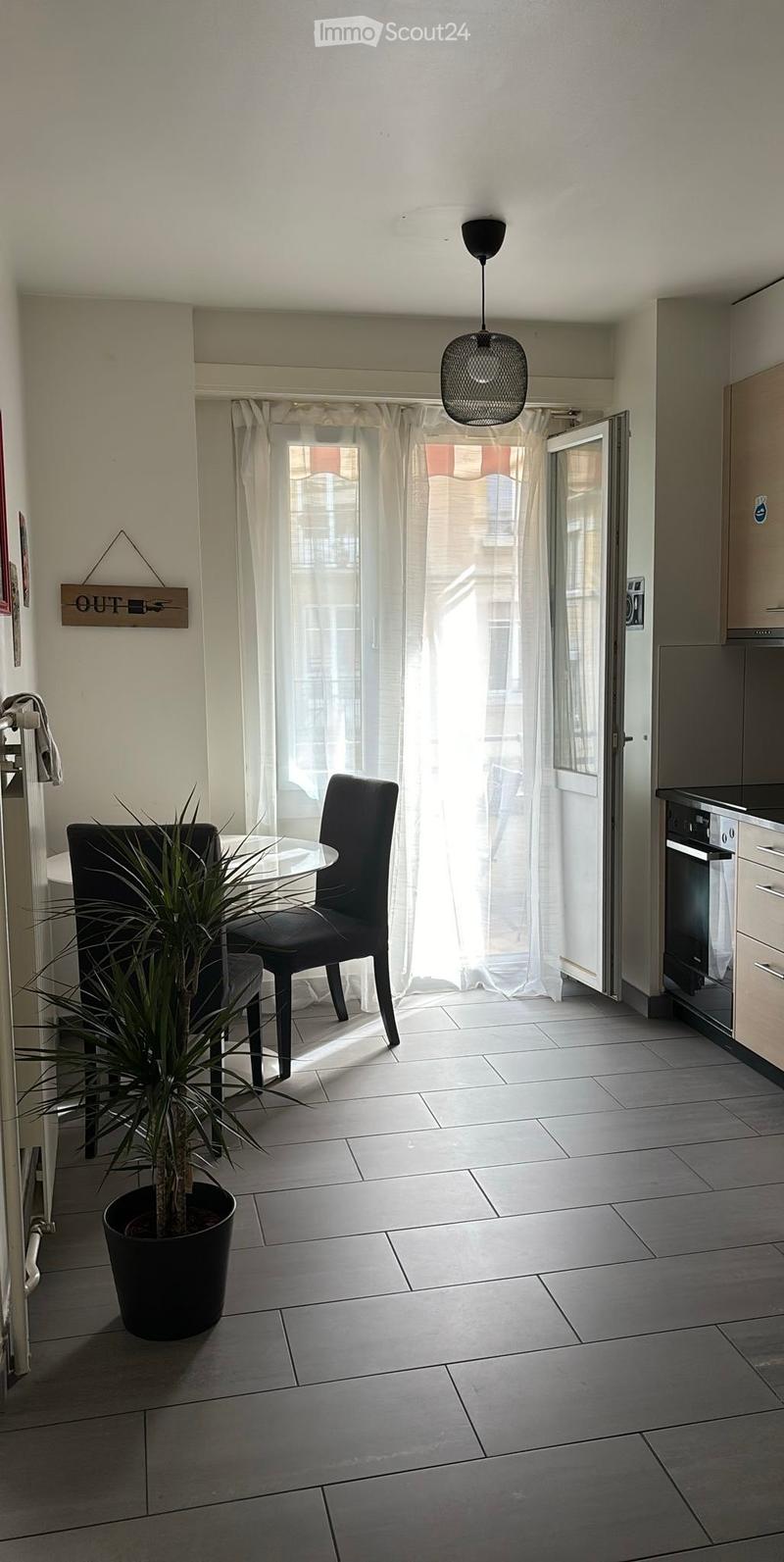 Modern, minimalist room with grey tiled flooring, white walls, and a large window with sheer curtains. Features a black hanging pendant light, a round glass table with two black chairs, a potted plant, and a kitchen area with cabinets and an oven.