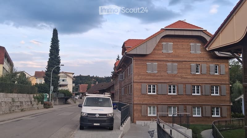 A two-story wooden house with a red-tiled roof and multiple windows, situated on a quiet street with a parked van nearby.