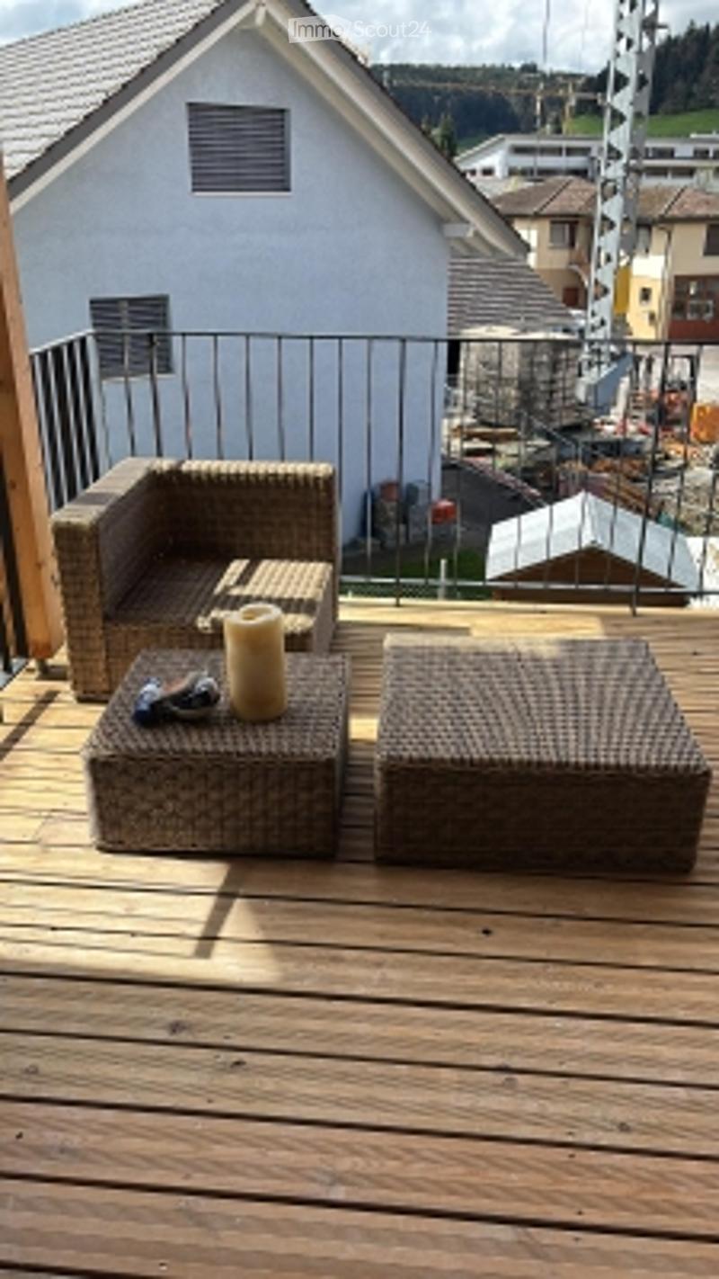 Wooden balcony with two wicker chairs and a small table, overlooking a residential area with houses and construction.