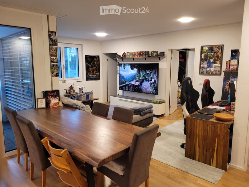 A modern, well-lit room with a wooden dining table and chairs, a sofa, a gaming chair, a desk with a laptop, and a large screen. Shelves with picture frames and decorative items are visible.