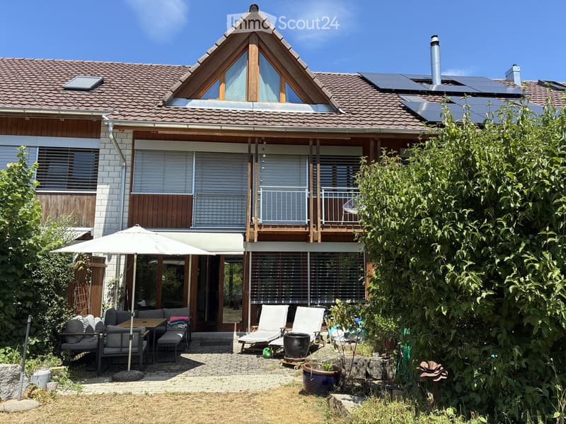 A modern two-story house with a well-maintained garden and patio area. The patio features a table, chairs, an umbrella, and lounge chairs. The house has large windows, a balcony, and solar panels on the roof.