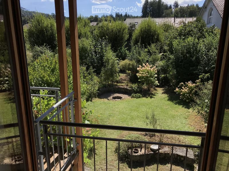 Balcony overlooking a well-maintained garden with grass, shrubs, and trees, with a stone fire pit and seating area visible below.