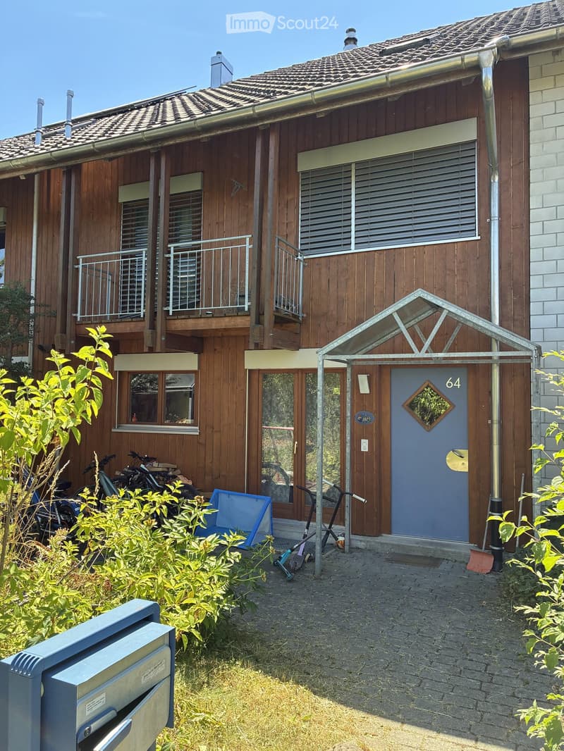 Two-story wooden house with a tiled roof, multiple windows, balconies, and a small entrance area with bicycles and a blue container.