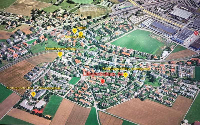 Aerial view of a residential area with labeled landmarks including Denner, Bahnhof RBS, Gemeindeverwaltung, Kindertagesstätte + Kindergarten, Längenbühlstr. 64, and Kindergarten, Real- + Sekundarschule. The area includes houses, green spaces, and a large