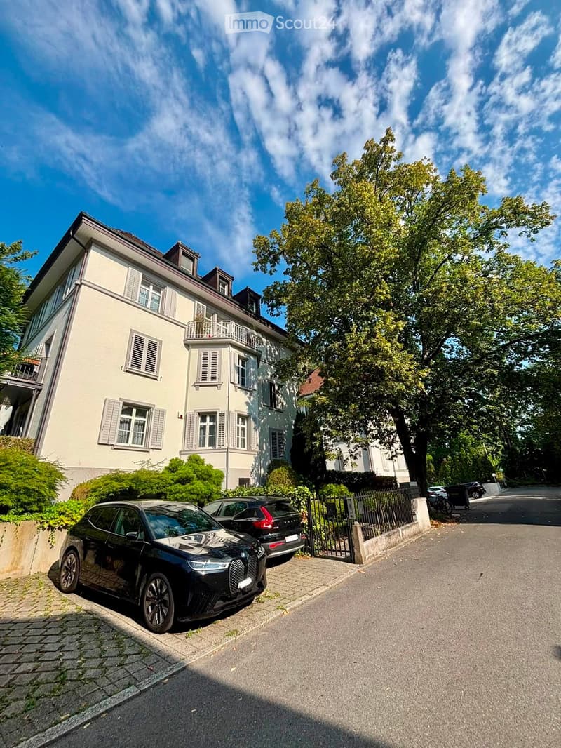 A multi-story residential house with light-colored walls, multiple windows with shutters, and a small balcony on the upper floor. The house is surrounded by greenery and has a gated driveway with parked cars.