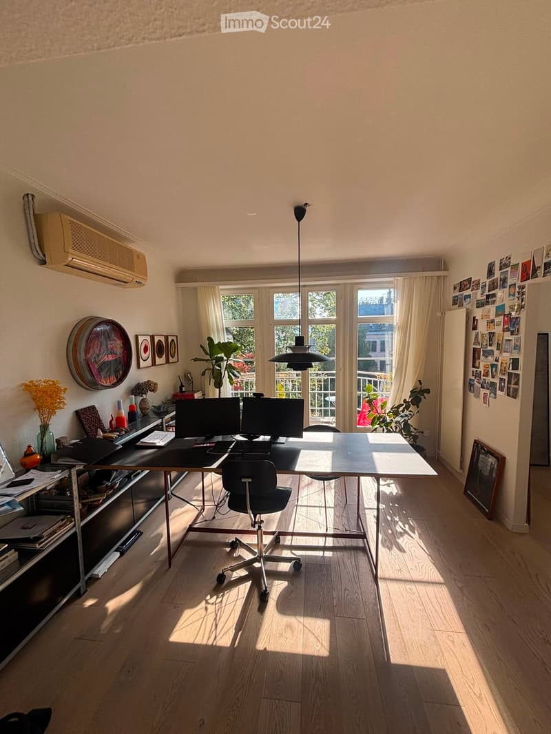 Bright, modern office space with wooden flooring, large windows, a desk with two monitors, a chair, bookshelf, and decorative items.