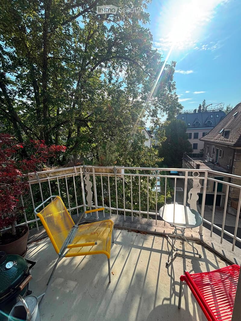 Balcony with a round glass table, yellow and red chairs, and a potted plant, overlooking a tree-lined street with buildings in the background.