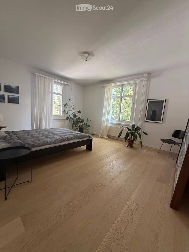 Bright bedroom with wooden flooring, large windows with white curtains, a bed with grey bedding, a small black chair, a round black table, and potted plants near the windows.