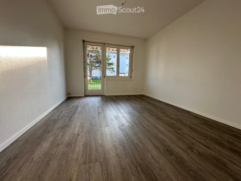 Empty room with wooden flooring, white walls, and large windows with sliding glass doors leading to an outdoor area.
