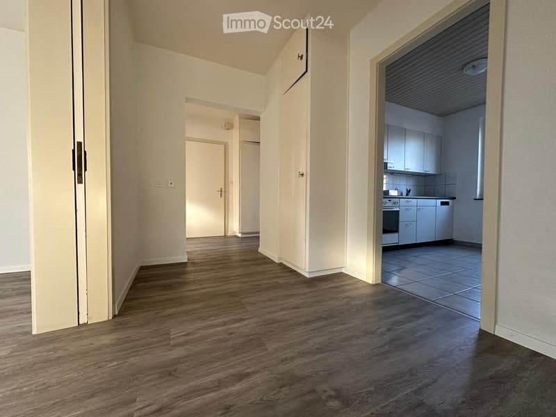 Empty hallway with wooden flooring, white walls, and doors leading to other rooms, including a kitchen with cabinets and appliances.