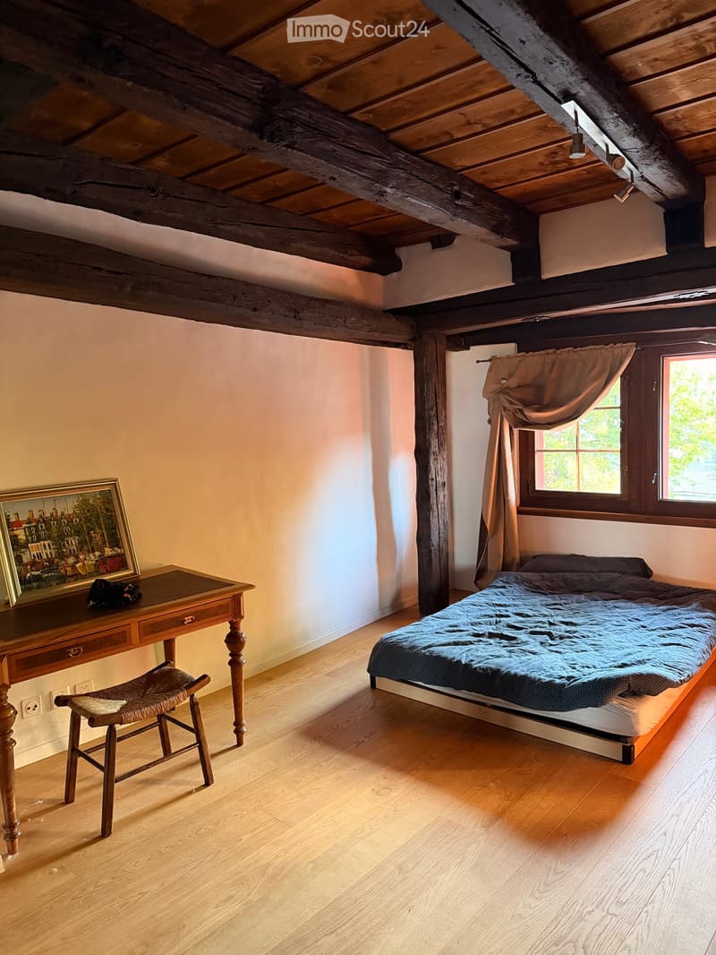 Cozy attic bedroom with wooden beams, a bed with blue bedding, a wooden desk with a chair, and a framed picture on the wall.