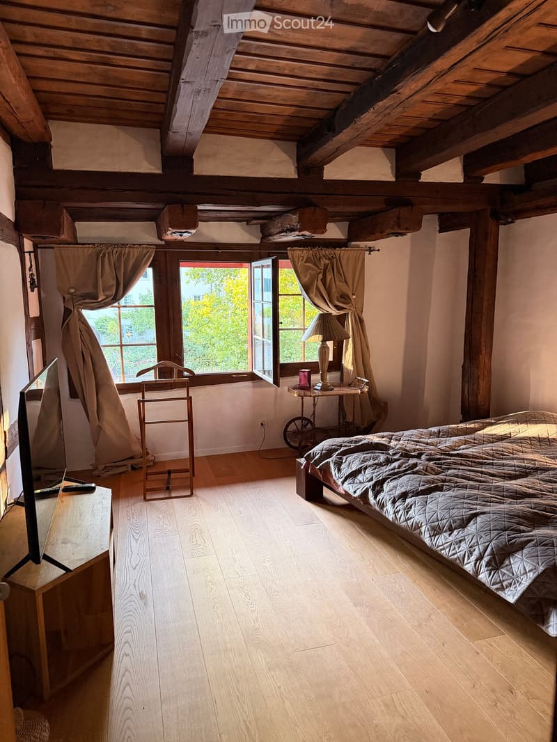 Cozy attic bedroom with wooden beams, large windows, a bed with a quilted cover, a small table with a lamp, and a chair near the window.