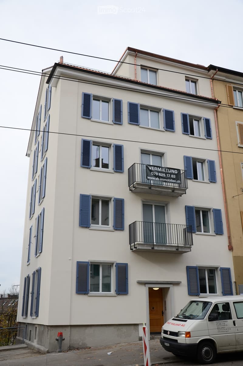 A three-story residential building with light beige walls, blue shutters, and multiple windows. Balconies are present on the upper floors, and a rental sign is displayed on one of the balconies. A white van is parked in front of the building.