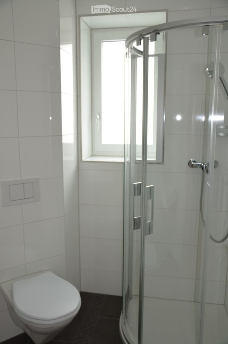 Modern bathroom with white tiled walls, a toilet, and a glass-enclosed shower. The shower has a sleek, contemporary design with silver handles and a built-in seat. The room features a small window above the shower for natural light.