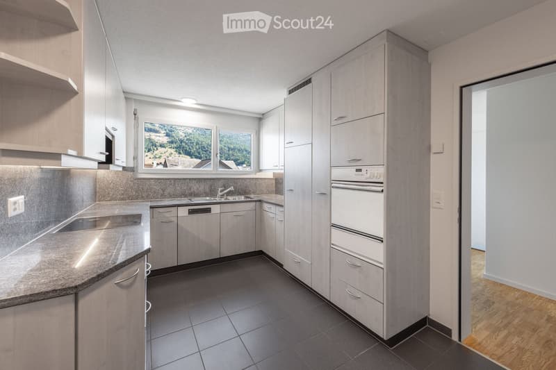 Modern kitchen with light wood cabinets, stainless steel sink, oven, and granite countertop. Large windows offer a scenic view.