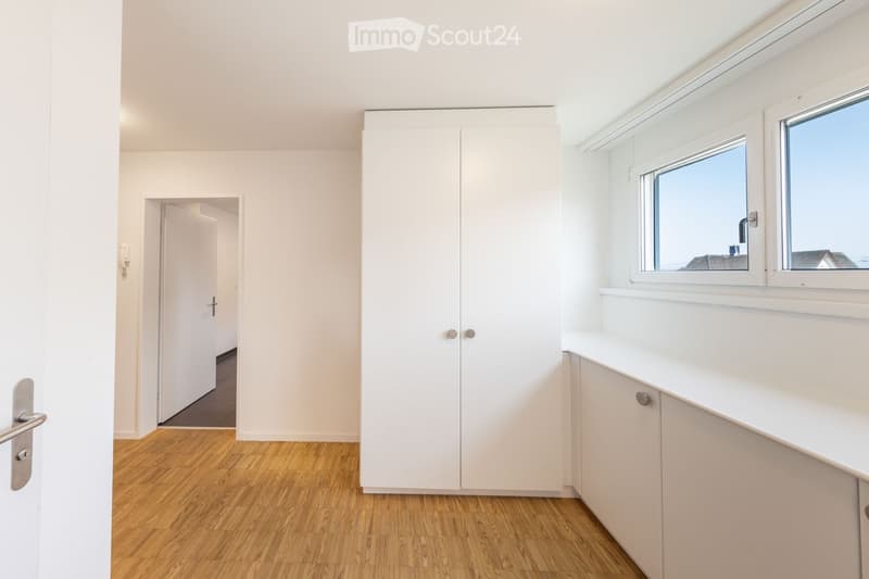 Empty room with white walls, wooden flooring, built-in white cabinetry, and large windows offering a view of the outside.