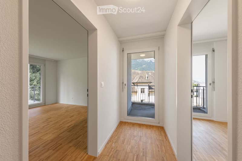 Bright, empty hallway with wooden flooring, white walls, and multiple doors leading to rooms and a balcony.