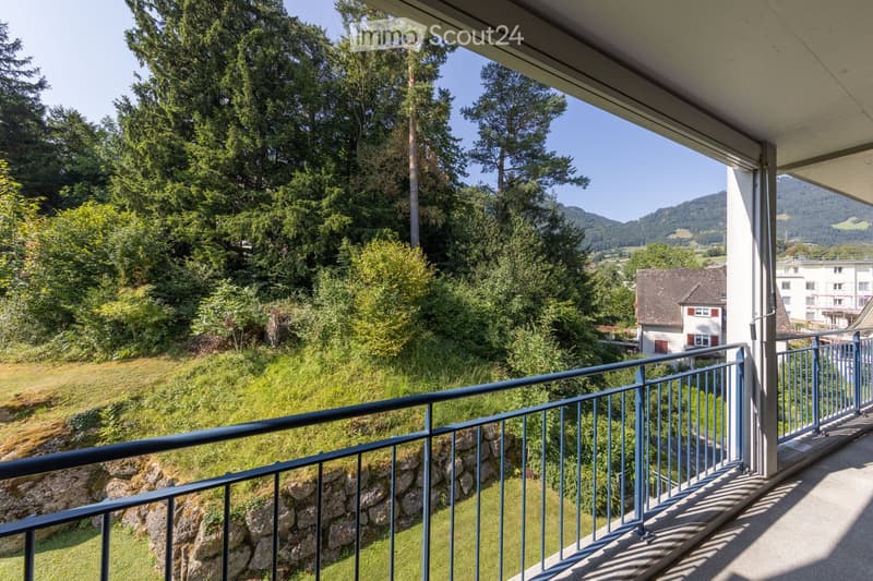 Balcony with metal railing overlooking a lush green landscape, trees, and distant mountains.