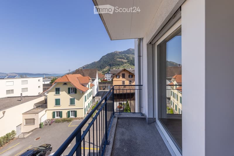 Balcony with a metal railing, overlooking a town with houses, a mountain, and a body of water in the distance.