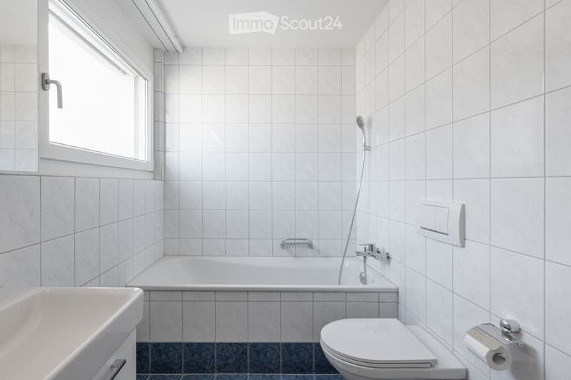 Modern bathroom with white tiled walls and floor, featuring a bathtub, toilet, sink, and showerhead.
