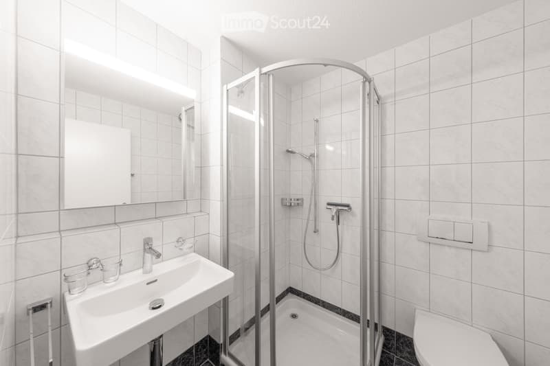 Modern bathroom with white tiled walls and floor, featuring a sink with a mirror, a glass-enclosed shower, and a toilet.
