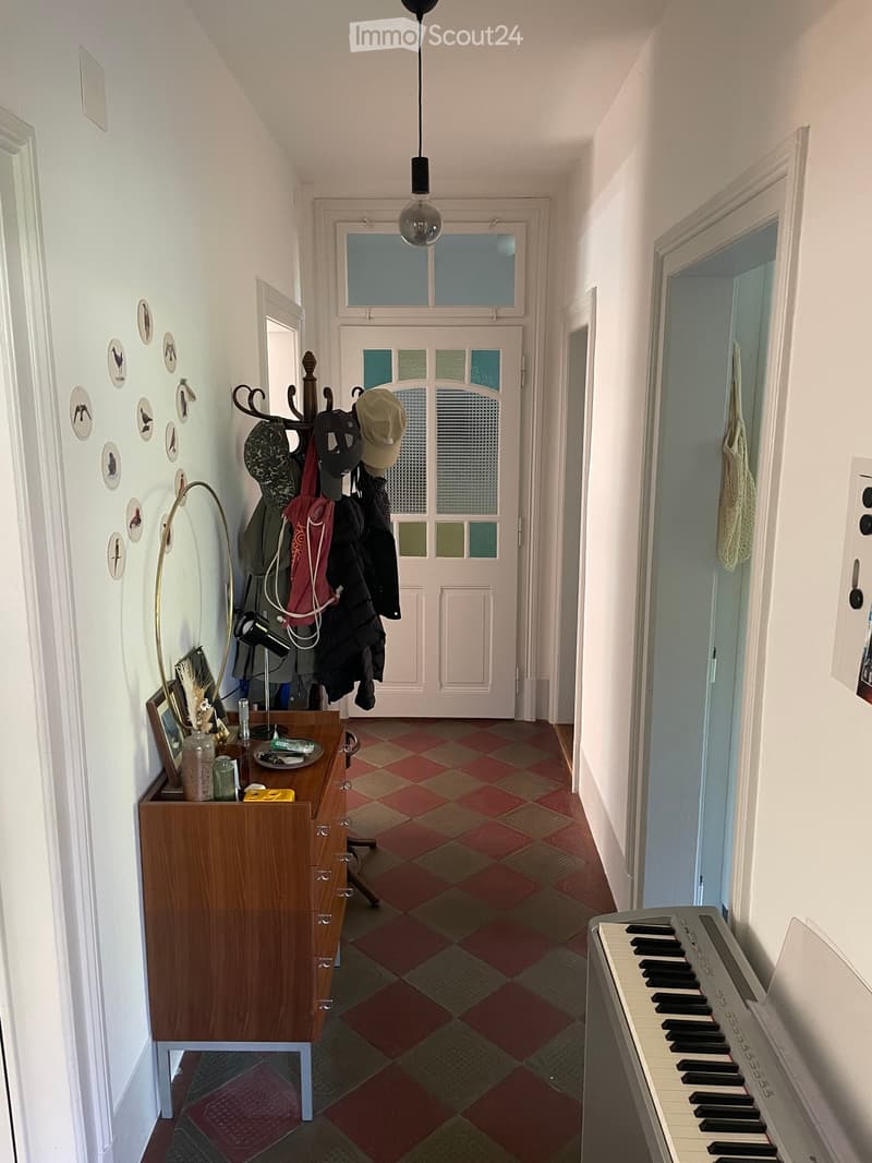 A narrow hallway with a checkered floor, white walls, and a hanging light fixture. A coat rack with jackets and hats is visible on the left, and a small wooden table with various items sits nearby. A piano is partially visible on the right side.