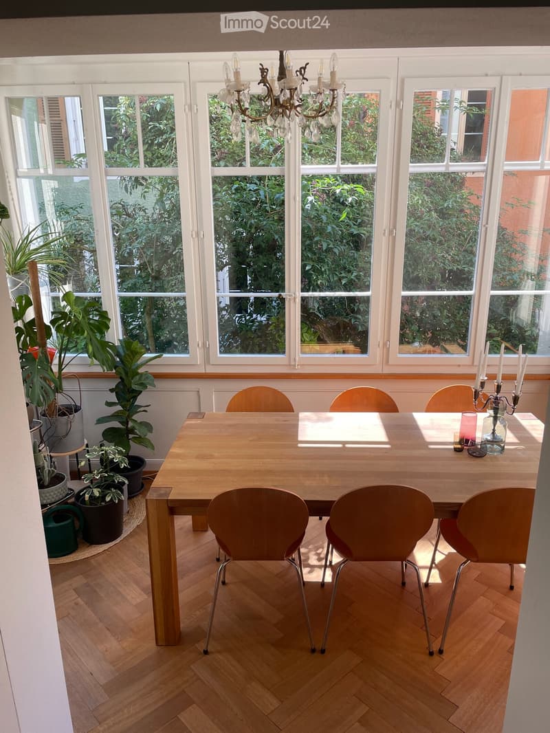Brightly lit dining room with large windows, wooden dining table, six chairs, candle holder with candles, and several potted plants.