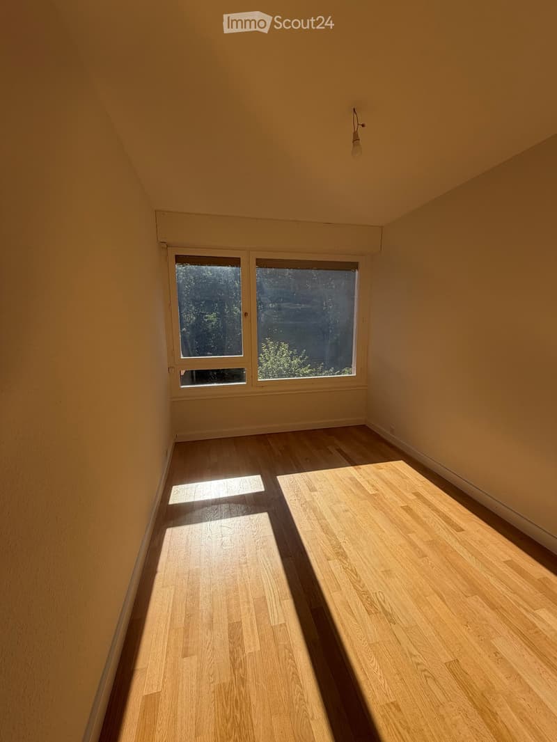 Empty room with light wooden flooring and large windows letting in natural light. The walls are painted a warm beige color, and there is a single light fixture hanging from the ceiling.