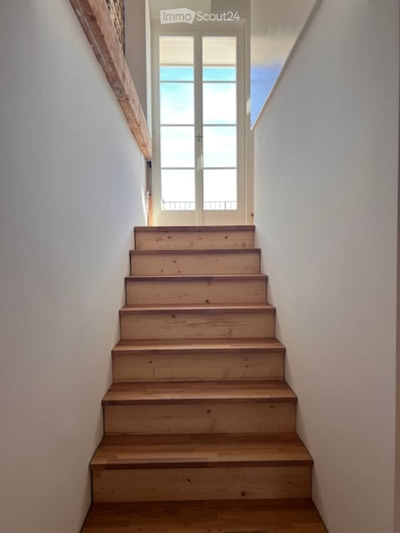 Wooden staircase leading up, white walls, a door at the top, and a window providing natural light.