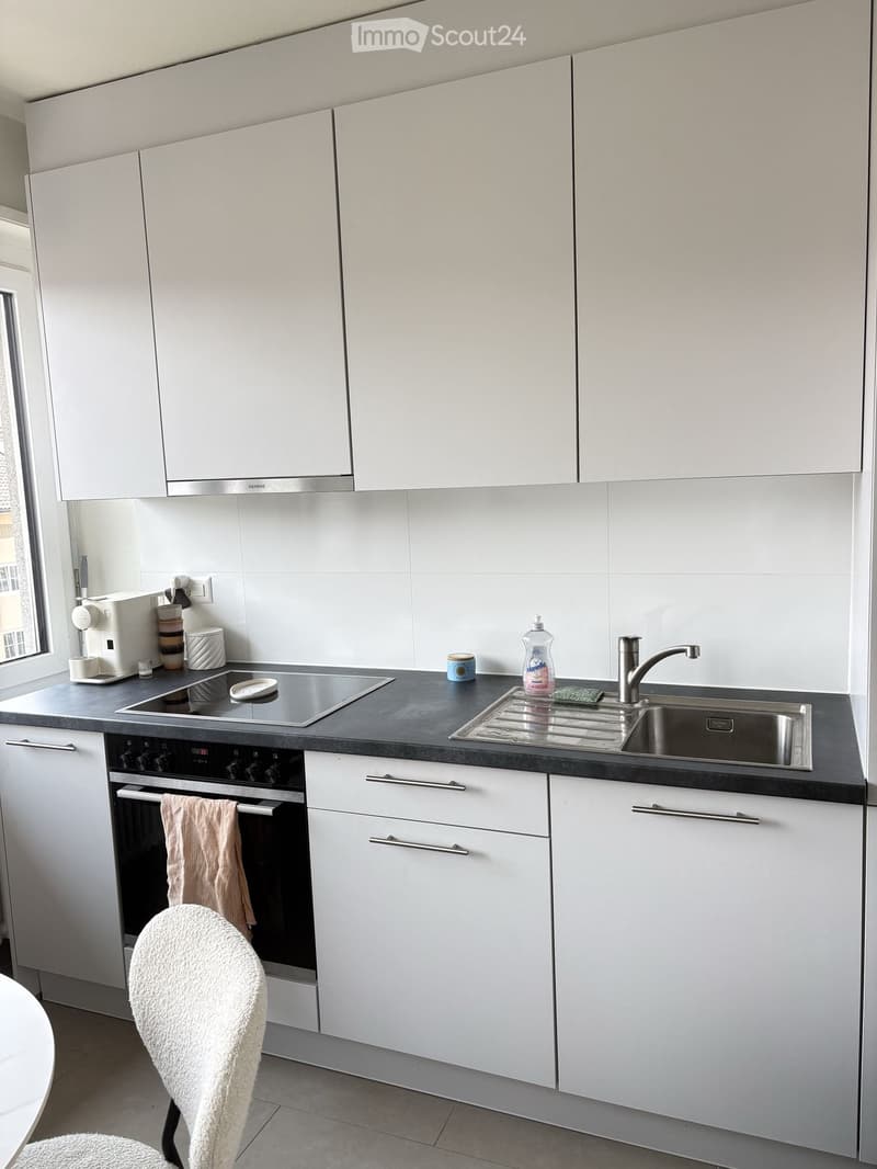 Modern kitchen with white cabinets, stainless steel appliances, and a black countertop. Features an oven, sink, faucet, and a white chair.
