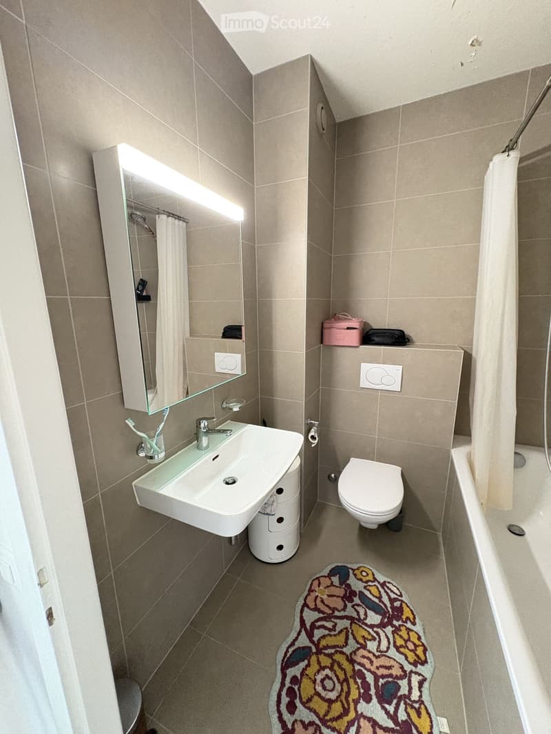 Modern bathroom with beige tiles, white sink, toilet, bathtub, and a floral rug on the floor.