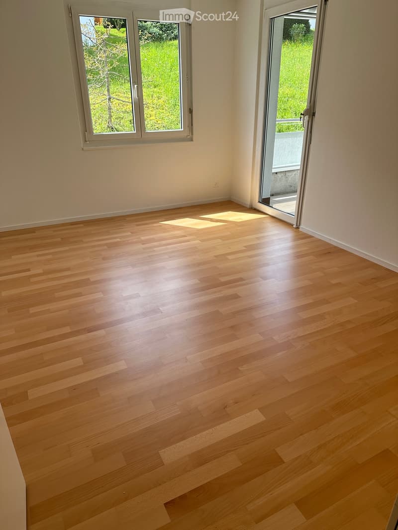 Empty room with light wooden flooring, white walls, and large windows with a view of greenery outside. A sliding glass door leads to a balcony.