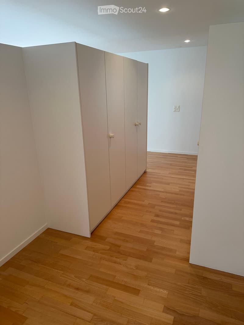 Empty hallway with light wooden flooring and white walls, featuring a set of closed white closet doors on one side.