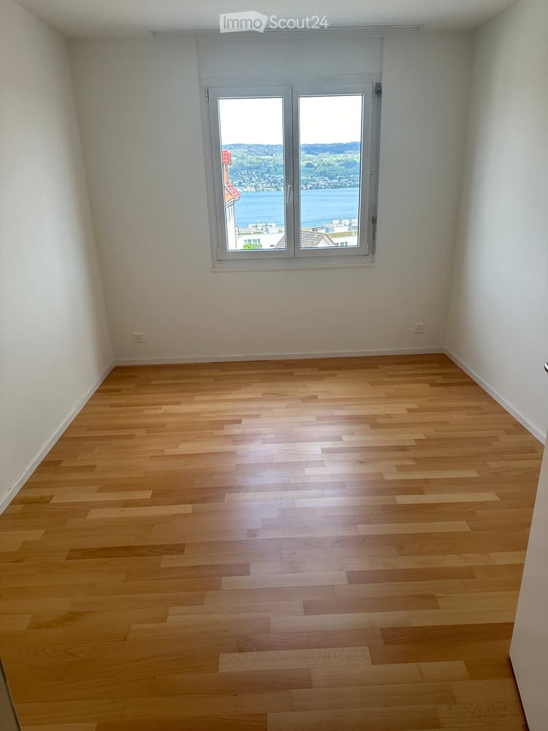 Empty room with light wooden flooring and white walls, featuring a large window with a scenic view of a lake and surrounding landscape.