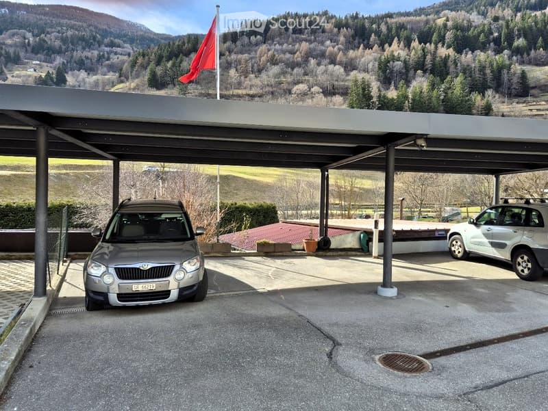 Carport with two parked cars, surrounded by a scenic mountainous landscape with trees and a flagpole.