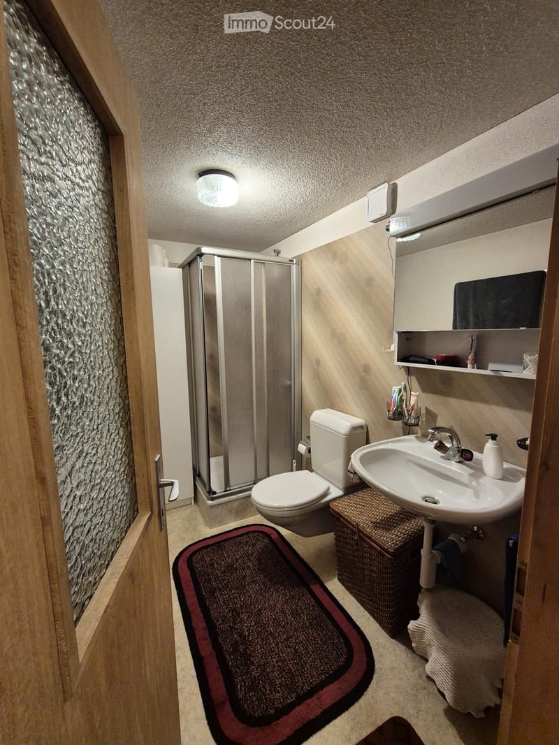 Modern bathroom with a toilet, sink, mirror, and glass-enclosed shower. Features a wicker basket, toothbrush holder, and a dark bath mat.