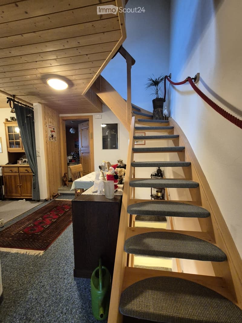 A cozy hallway with wooden beams, a staircase with carpeted steps, a small table with a Santa decoration, a green watering can, and a cabinet with glass doors.