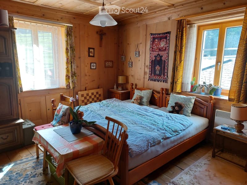 Cozy wooden bedroom with a double bed, nightstands, a small table with a plant, and wooden chairs. The room features wooden walls, a hanging light fixture, and decorative elements like a tapestry and framed pictures.