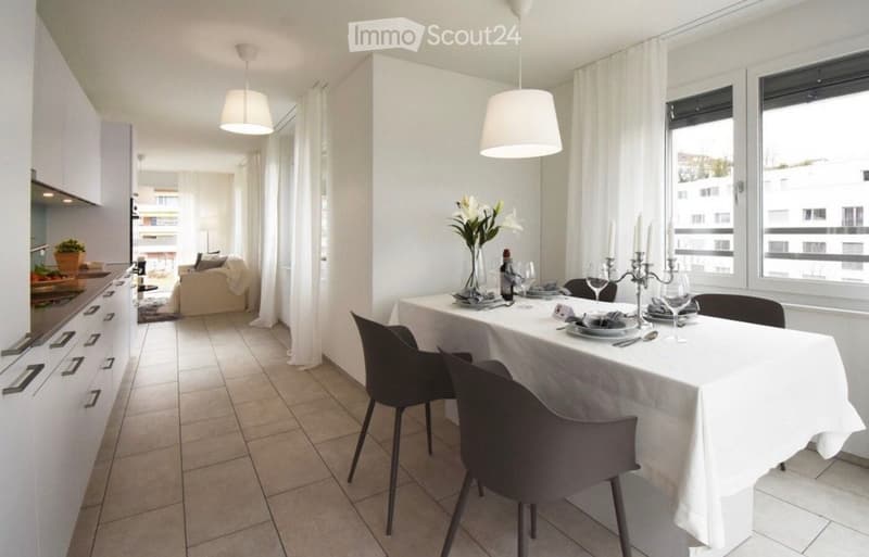 Modern open-plan living and dining area with white walls, tiled floor, and large windows. Features a dining table with chairs, a sofa, kitchen cabinets, and decorative items.