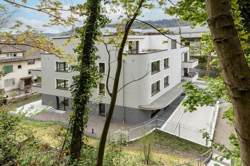 Modern, multi-story house with white exterior, large windows, and a balcony. The house is surrounded by trees and greenery, with a raised ground floor and a view of the surrounding landscape.