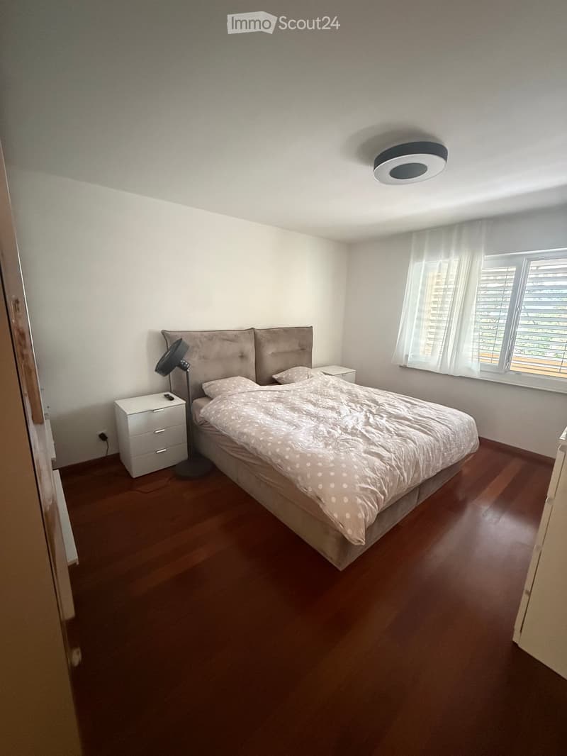 Modern bedroom with wooden flooring, a large bed with a tufted headboard, nightstands, and a lamp. Large window with blinds and sheer curtains.