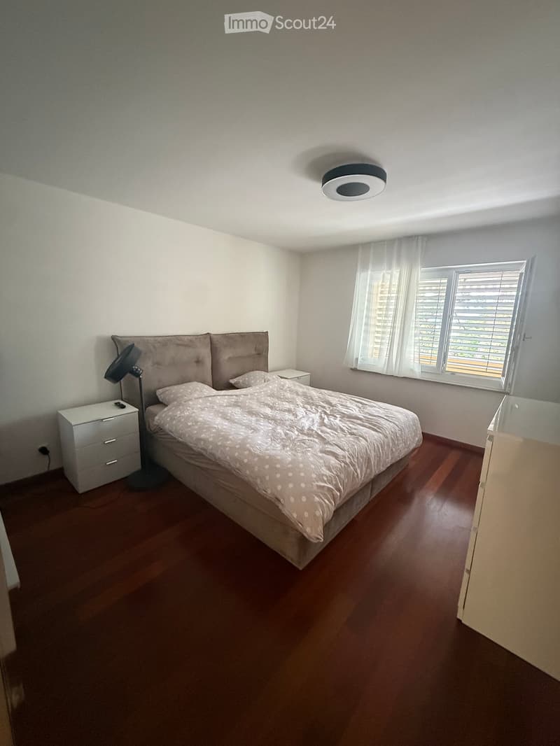 A well-lit bedroom with a wooden floor, a large bed with a patterned duvet, a nightstand, a lamp, and a drawer unit.