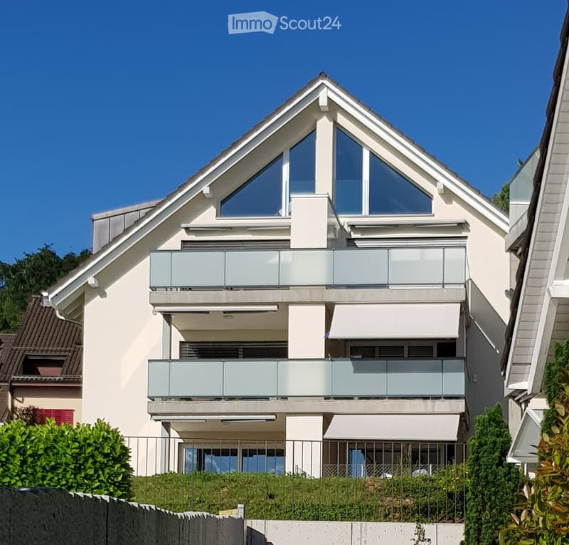 Modern two-story house with white exterior, large windows, and balconies. Features a triangular roof and contemporary design.