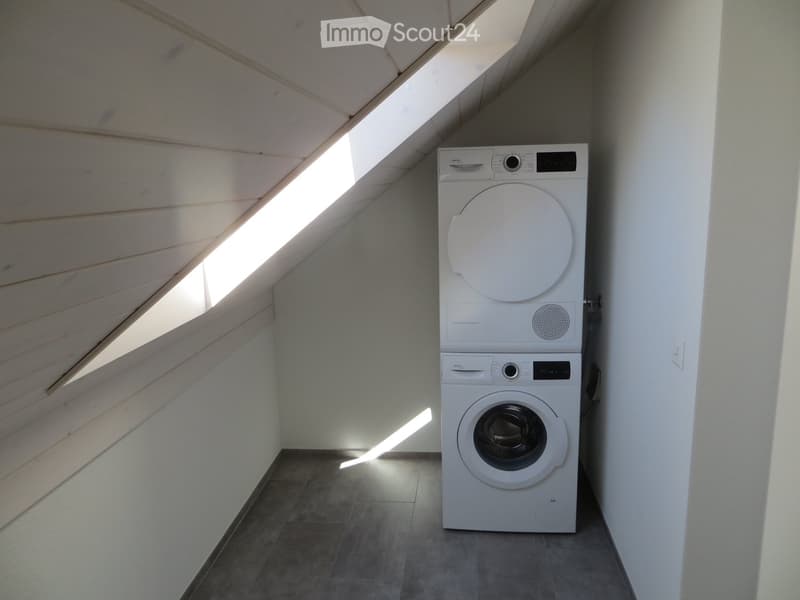 A small, well-lit room with a slanted wooden ceiling and a skylight, containing a stacked washing machine and dryer.