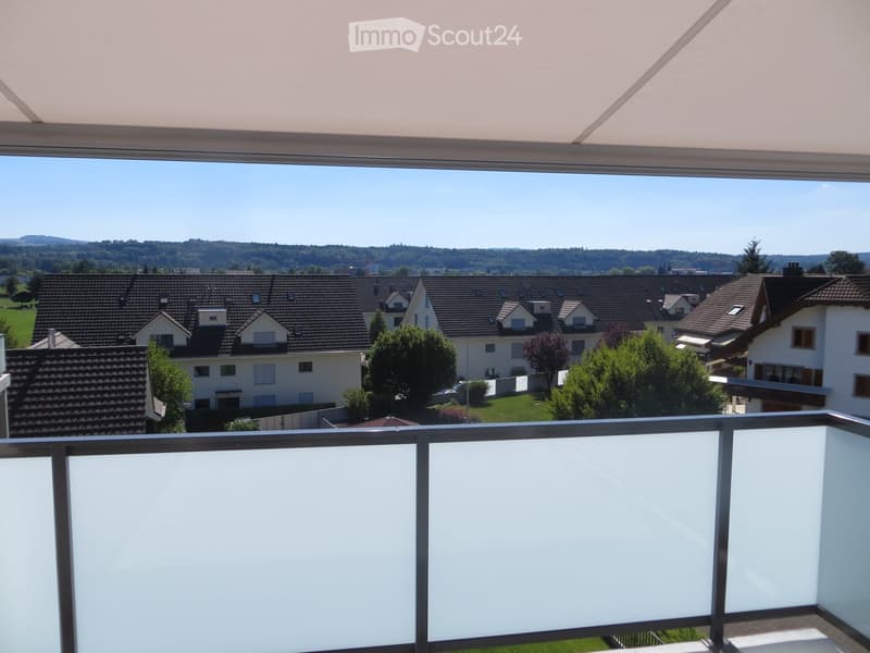 Balcony with a view of a suburban neighborhood, featuring houses with pitched roofs, trees, and distant hills under a clear blue sky.
