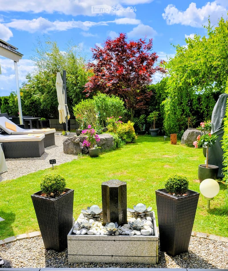 Well-maintained garden with a central water fountain, surrounded by potted plants, lounge chairs, and a variety of trees and shrubs.