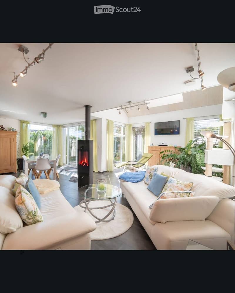Modern living room with white sofas, colorful cushions, a glass coffee table, a central fireplace, large windows with yellow curtains, and a dining area with chairs and a table.