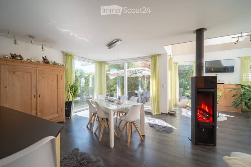 Bright, open-plan living and dining area with large windows, a modern fireplace, a dining table with chairs, a wooden cabinet, and a television mounted on the wall.