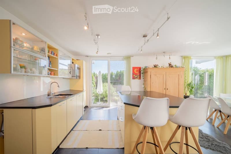Modern kitchen with white and yellow cabinets, a central island, bar stools, and a dining area with chairs and a table. The space features a sink, a microwave, and ample natural light from large windows.