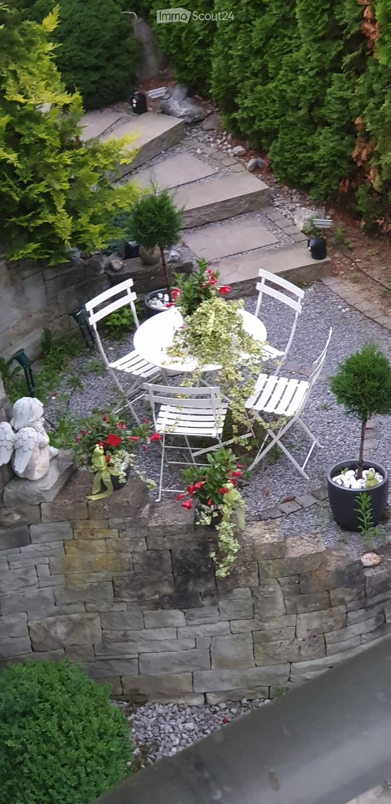A small, well-maintained garden area with a stone wall, a round white table surrounded by four white chairs, potted plants, and a decorative statue.